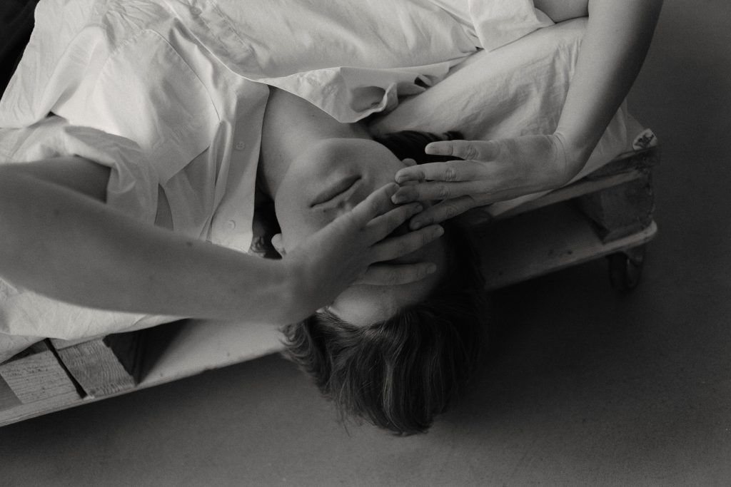 Artistic black and white photo of a brunette woman with hands on face, lying on a wooden pallet.
